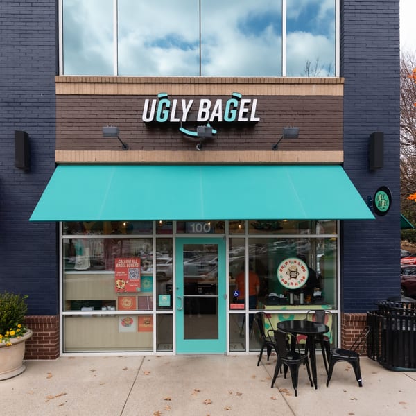 The exterior of Ugly Bagel, featuring a teal awning, black outdoor seating, and brick facade.