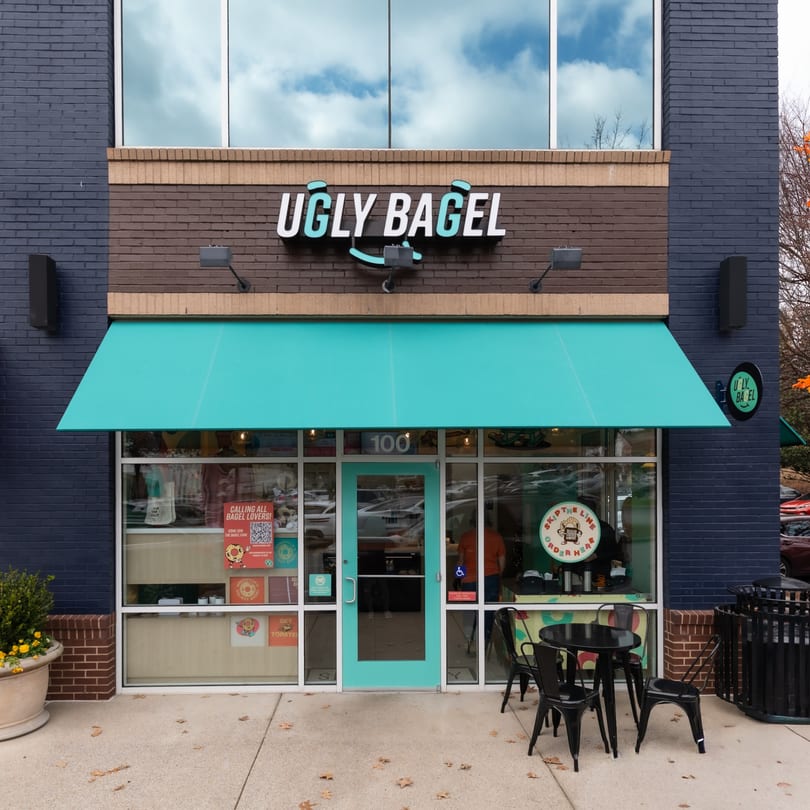 The exterior of Ugly Bagel, featuring a teal awning, black outdoor seating, and brick facade.