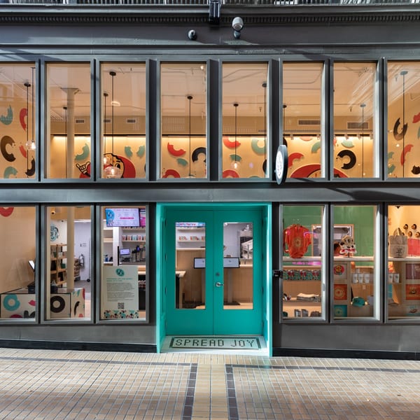 Exterior of Ugly Bagel shop with a bright teal door, large windows, and "SPREAD JOY" on the mat.